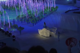 A vibrant performance scene with performers wearing costumes that resemble green leaves. The setting is illuminated with blue and purple lights, giving it a magical, ethereal feel. At the forefront, a person is dressed in a flowing gown adorned with lights, adding a touch of elegance to the scene.