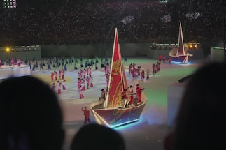 A large indoor event featuring performers wearing colorful costumes, predominantly in pink, with illuminated boat-like structures on the stage. The audience is in the background, as seen from a distance, with dim lighting and spotlight effects highlighting the performers.