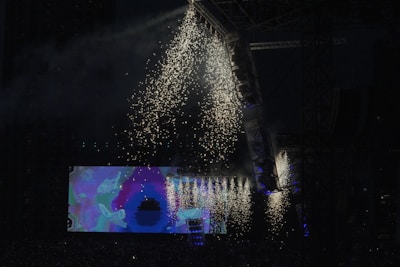 A vibrant display of fireworks illuminates an outdoor concert stage during a night event. Bright sparks cascade from the top of a large stage structure, highlighting the darkness surrounding it. On the stage, a large video screen displays colorful abstract visuals, adding to the atmosphere of the event.