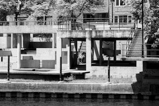 A black-and-white photograph capturing a concrete urban structure with geometric shapes and shadows. The scene includes staircases, railings, and trees in the background. A person is seated in the center of the structure, adding an element of human presence amid the architectural forms.