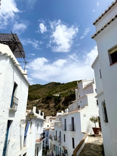 A narrow street flanked by white-washed buildings with small balconies, set against a backdrop of hills. The architecture reflects a traditional Mediterranean style, with bright blue sky and scattered clouds above, contributing to a peaceful and serene atmosphere.