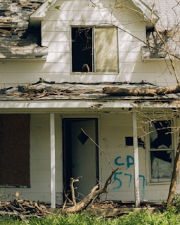 The image depicts an abandoned house with a dilapidated exterior. The roof is partially damaged and there are branches and debris scattered across the porch. One of the upstairs windows has a large bird perched on the sill. The lower floor has broken windows and graffiti with the letters 'CP 577' written in blue spray paint. The surrounding greenery suggests the house is in a neglected area.