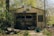 A small, abandoned garage with a wooden structure is overgrown with vegetation and covered in graffiti. The garage door is closed, and there is a lot of shrubbery surrounding the building. Various debris, including chopped wood and discarded items, is scattered in the foreground, giving the area a neglected and forgotten appearance.