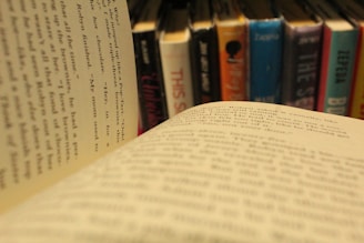 A close-up view of an open book with text visible on the pages, set against a blurred background of vertically stacked books on a shelf. The image focuses on the text, creating an impression of reading or studying in a library or bookstore.