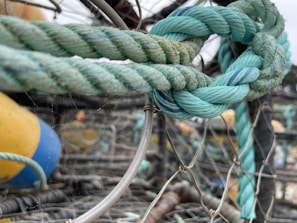 A close-up of a thick, intertwined rope with shades of teal and blue. The knot is tied securely among metal wires and fishing nets. In the background, a yellow and blue buoy is slightly out of focus, giving a nautical setting.