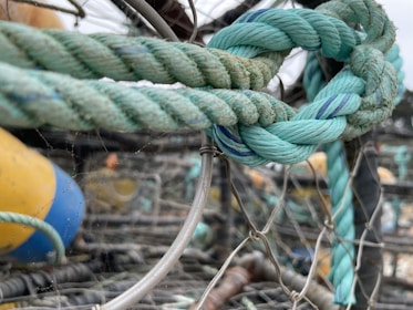 A close-up of a thick, intertwined rope with shades of teal and blue. The knot is tied securely among metal wires and fishing nets. In the background, a yellow and blue buoy is slightly out of focus, giving a nautical setting.