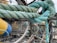 A close-up of a thick, intertwined rope with shades of teal and blue. The knot is tied securely among metal wires and fishing nets. In the background, a yellow and blue buoy is slightly out of focus, giving a nautical setting.