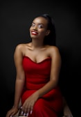A person wearing a strapless red dress is sitting against a dark background. The individual has a confident and warm smile, with their hands resting on their lap.