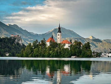 A serene lake with a small island at its center, featuring a picturesque church with a prominent bell tower. The island is lush with greenery, surrounded by calm waters that mirror the landscape. In the background, a range of mountainous peaks stretches across the horizon under a partly cloudy sky. Another building is visible atop a cliff to the left of the island.