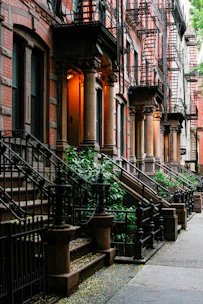 A row of classic brownstone buildings is seen with ornate columns flanking each entryway, characteristic black wrought iron railings, and external staircases leading up to wooden doors. Lush green plants grow along the sidewalk, adding a touch of nature to the urban setting. Fire escapes are visible on the facades of the buildings.