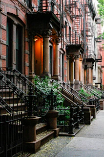 A row of classic brownstone buildings is seen with ornate columns flanking each entryway, characteristic black wrought iron railings, and external staircases leading up to wooden doors. Lush green plants grow along the sidewalk, adding a touch of nature to the urban setting. Fire escapes are visible on the facades of the buildings.