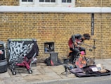 A street musician with a unique setup is performing against a brick wall. The artist is playing a double-neck guitar while surrounded by various musical equipment, including an amplifier and a microphone. Nearby, a stroller and some clothing are placed next to a graffiti-covered utility box.