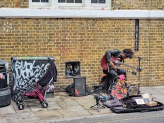 A street musician with a unique setup is performing against a brick wall. The artist is playing a double-neck guitar while surrounded by various musical equipment, including an amplifier and a microphone. Nearby, a stroller and some clothing are placed next to a graffiti-covered utility box.