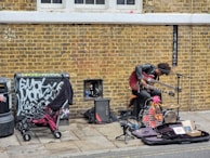 A street musician with a unique setup is performing against a brick wall. The artist is playing a double-neck guitar while surrounded by various musical equipment, including an amplifier and a microphone. Nearby, a stroller and some clothing are placed next to a graffiti-covered utility box.