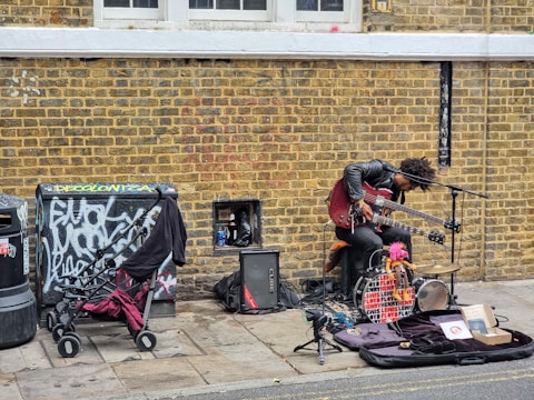 A street musician with a unique setup is performing against a brick wall. The artist is playing a double-neck guitar while surrounded by various musical equipment, including an amplifier and a microphone. Nearby, a stroller and some clothing are placed next to a graffiti-covered utility box.