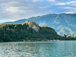 A picturesque lake with calm waters reflecting the surrounding scenery. In the background, a rocky hill rises sharply with a historic castle perched on top. The hill is covered with dense green forest, and behind it, majestic mountains are partially covered by soft clouds. The sky is blue with patches of clouds, providing a serene backdrop.