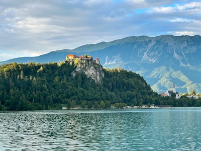 A picturesque lake with calm waters reflecting the surrounding scenery. In the background, a rocky hill rises sharply with a historic castle perched on top. The hill is covered with dense green forest, and behind it, majestic mountains are partially covered by soft clouds. The sky is blue with patches of clouds, providing a serene backdrop.