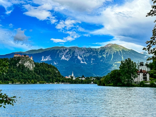 A scenic landscape with a large, calm lake in the foreground, surrounded by lush green trees. To the left, a historic castle is perched on a rocky cliff overlooking the water. In the background, majestic mountains rise under a partly cloudy blue sky, adding to the grandeur of the scene. A small white church with a steeple is visible near the lake.