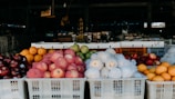 A market stall displaying a variety of fruits stacked in white plastic crates. There are apples wrapped in pink nets, white mesh-covered pears, oranges, and apples with dark red skins. The background is filled with various indistinct items suggesting a bustling market environment.