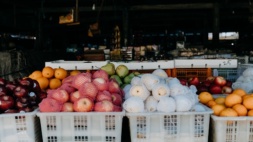 A market stall displaying a variety of fruits stacked in white plastic crates. There are apples wrapped in pink nets, white mesh-covered pears, oranges, and apples with dark red skins. The background is filled with various indistinct items suggesting a bustling market environment.