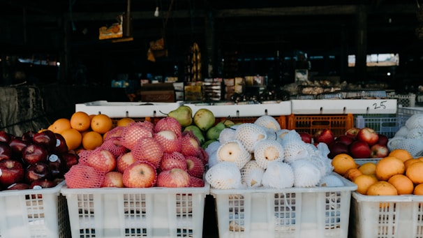 A market stall displaying a variety of fruits stacked in white plastic crates. There are apples wrapped in pink nets, white mesh-covered pears, oranges, and apples with dark red skins. The background is filled with various indistinct items suggesting a bustling market environment.