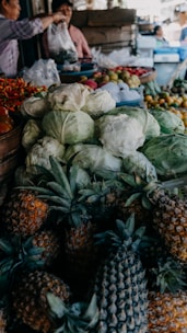 A bustling market stall filled with fresh produce, including piles of cabbages, pineapples, and various fruits like apples and pomegranates. In the background, two people are engaged in a transaction, with one person handing over a plastic bag. The scene is lively with multiple items stacked closely together.