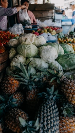 A bustling market stall filled with fresh produce, including piles of cabbages, pineapples, and various fruits like apples and pomegranates. In the background, two people are engaged in a transaction, with one person handing over a plastic bag. The scene is lively with multiple items stacked closely together.