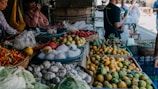 A busy market scene showcasing a variety of fresh produce including garlic, chilies, oranges, apples, and other vegetables. Several people are engaged in conversation and browsing the stalls. The setting is vibrant and bustling, filled with baskets and containers overflowing with colorful fruits and vegetables.