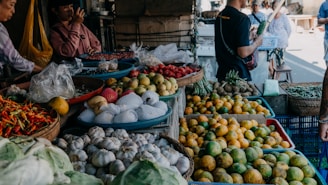 A busy market scene showcasing a variety of fresh produce including garlic, chilies, oranges, apples, and other vegetables. Several people are engaged in conversation and browsing the stalls. The setting is vibrant and bustling, filled with baskets and containers overflowing with colorful fruits and vegetables.