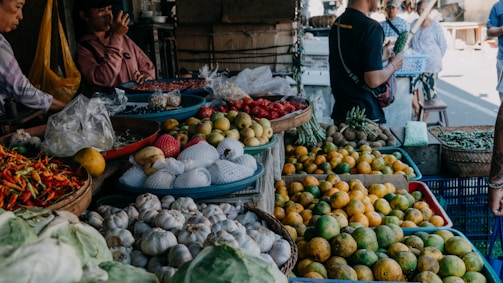 A busy market scene showcasing a variety of fresh produce including garlic, chilies, oranges, apples, and other vegetables. Several people are engaged in conversation and browsing the stalls. The setting is vibrant and bustling, filled with baskets and containers overflowing with colorful fruits and vegetables.