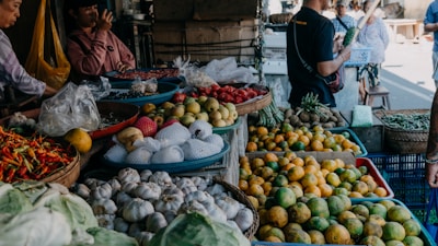 A busy market scene showcasing a variety of fresh produce including garlic, chilies, oranges, apples, and other vegetables. Several people are engaged in conversation and browsing the stalls. The setting is vibrant and bustling, filled with baskets and containers overflowing with colorful fruits and vegetables.
