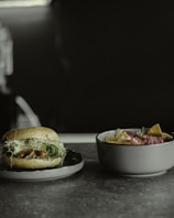 A close-up of a sandwich on a plate with fresh ingredients such as leafy greens, sprouts, and cucumber slices. Next to it is a bowl filled with tortilla chips and topped with sliced meat and herbs.