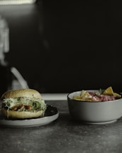A close-up of a sandwich on a plate with fresh ingredients such as leafy greens, sprouts, and cucumber slices. Next to it is a bowl filled with tortilla chips and topped with sliced meat and herbs.