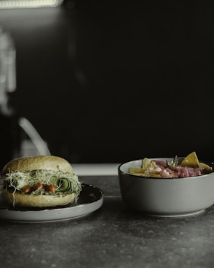 A close-up of a sandwich on a plate with fresh ingredients such as leafy greens, sprouts, and cucumber slices. Next to it is a bowl filled with tortilla chips and topped with sliced meat and herbs.