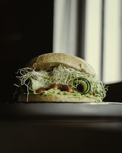 A sandwich is artfully assembled with layers of fresh ingredients including sprouts, cucumber slices, and a green spread, possibly avocado, between two slices of bread. The composition sits elegantly on a reflective surface, with the background softly blurred, suggesting a natural, possibly morning lighting setup.