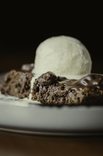 A close-up view of a chocolate dessert topped with a scoop of vanilla ice cream. The dessert appears to be a moist brownie or cake with a shiny chocolate glaze. The creamy ice cream is beginning to melt slightly over the warm dessert, creating a delectable combination.