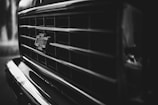 A close-up, black and white image of a vehicle's front grille with a Chevrolet logo prominently displayed. The grille has a grid pattern and the image has a moody and metallic texture.