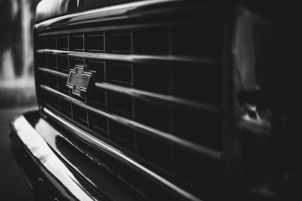 A close-up, black and white image of a vehicle's front grille with a Chevrolet logo prominently displayed. The grille has a grid pattern and the image has a moody and metallic texture.