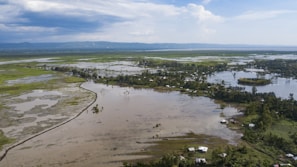 An aerial view of a large flooded area with scattered houses, surrounded by lush greenery and patches of waterlogged land. The landscape stretches towards distant hills under a partly cloudy sky.