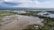 An aerial view of a large flooded area with scattered houses, surrounded by lush greenery and patches of waterlogged land. The landscape stretches towards distant hills under a partly cloudy sky.