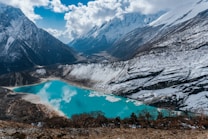 A stunning alpine scene features a tranquil turquoise lake surrounded by snow-covered mountains. The rugged peaks rise sharply against a partly cloudy sky, reflecting on the lake's surface. Sparse vegetation can be seen around the rocky terrain, adding a rich texture to the landscape.