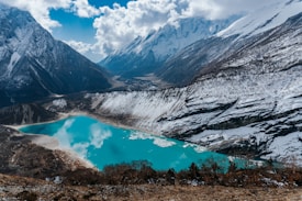 A stunning alpine scene features a tranquil turquoise lake surrounded by snow-covered mountains. The rugged peaks rise sharply against a partly cloudy sky, reflecting on the lake's surface. Sparse vegetation can be seen around the rocky terrain, adding a rich texture to the landscape.