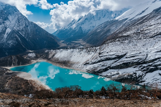 A stunning alpine scene features a tranquil turquoise lake surrounded by snow-covered mountains. The rugged peaks rise sharply against a partly cloudy sky, reflecting on the lake's surface. Sparse vegetation can be seen around the rocky terrain, adding a rich texture to the landscape.