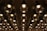 A ceiling installation composed of numerous circular, transparent glass light fixtures with metallic supports. The fixtures are arranged in a grid pattern, reflecting ambient light and casting soft glows. The reflection on the mirrored surface enhances the symmetrical appearance.