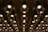 A ceiling installation composed of numerous circular, transparent glass light fixtures with metallic supports. The fixtures are arranged in a grid pattern, reflecting ambient light and casting soft glows. The reflection on the mirrored surface enhances the symmetrical appearance.