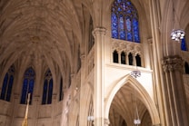 The photograph captures the intricate interior of a cathedral, focusing on the soaring stone arches and detailed Gothic architecture. Stained glass windows with vibrant blue hues are prominently featured, casting a colorful illumination across the stone surfaces. Hanging chandeliers add to the ornate design, reflecting the grandeur and artistry of the space.