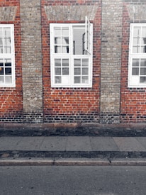 A brick wall with evenly spaced windows, each featuring white frames. The windows have multiple panes, and one is partially open. The wall consists of reddish-brown and grey bricks, with distinct texture and pattern. A sidewalk runs along the base of the building, made of gray stone slabs and cobblestones.