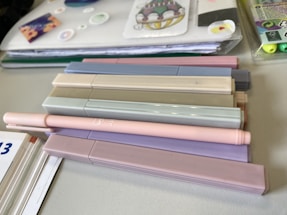 A collection of pastel-colored markers is arranged on a table alongside a notebook with decorative stickers and papers. The markers are mostly in soft shades of beige, pink, gray, and purple. Next to the markers, there is a visible edge of a calendar or workbook and some highlighter pens in a plastic case.