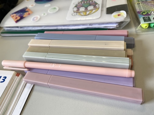 A collection of pastel-colored markers is arranged on a table alongside a notebook with decorative stickers and papers. The markers are mostly in soft shades of beige, pink, gray, and purple. Next to the markers, there is a visible edge of a calendar or workbook and some highlighter pens in a plastic case.