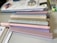 A collection of pastel-colored markers is arranged on a table alongside a notebook with decorative stickers and papers. The markers are mostly in soft shades of beige, pink, gray, and purple. Next to the markers, there is a visible edge of a calendar or workbook and some highlighter pens in a plastic case.